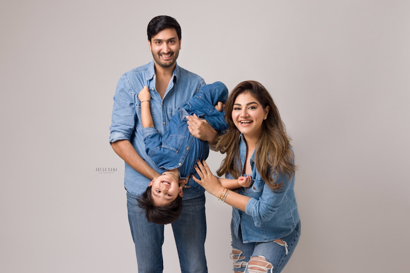 Dad playing with his son while mom poses cutely, all wearing denims against a grey backdrop, captured during a family photography session by Anega Bawa in Delhi, Gurgaon. Their joyful interactions highlight the warmth and fun of family life.