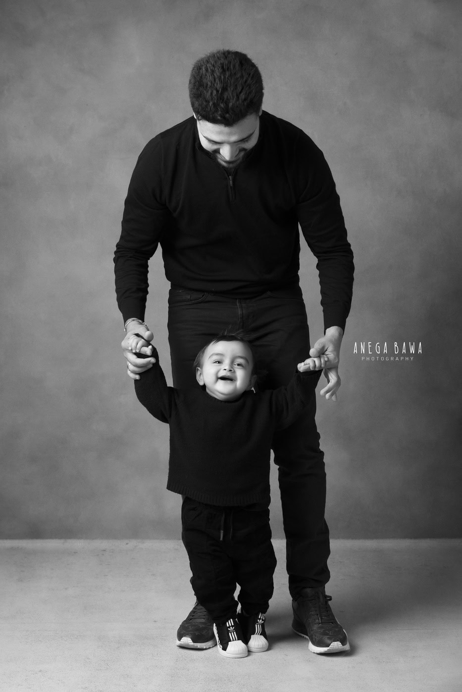 Dad and son twinning in black, standing together against a grey backdrop, captured during a family photography session by Anega Bawa in Delhi, Gurgaon. Their coordinated outfits and joyful expressions highlight their strong connection.