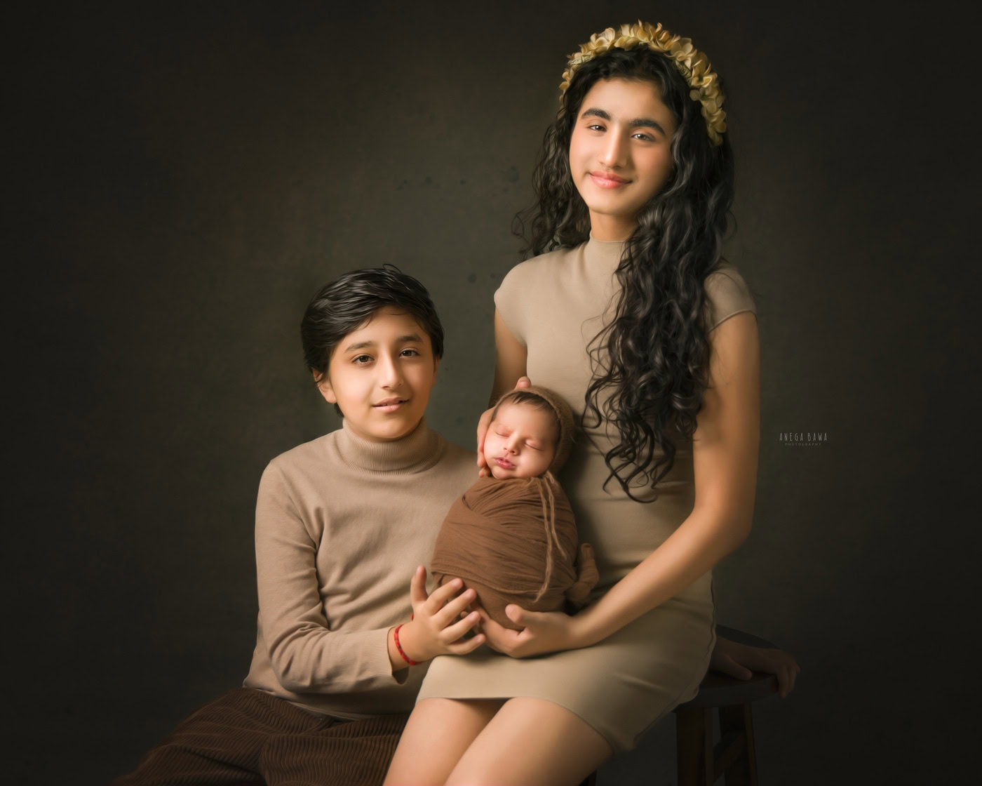 Elder sister holding her newborn brother wrapped in a brown wrap, posing with their elder brother in a beige dress against a brown backdrop during a family photography session by Anega Bawa in Delhi, Gurgaon. Their warm smiles and close-knit interaction beautifully illustrate the bond of sibling love.