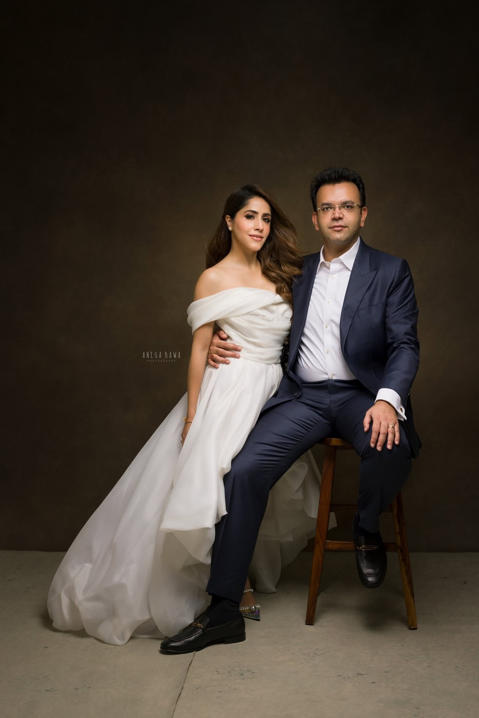 Husband sitting on a stool, posing with his wife in a white gown against a brown backdrop, captured during a family photography session by Anega Bawa in Delhi, Gurgaon. Their elegant poses and warm expressions showcase their love and connection.