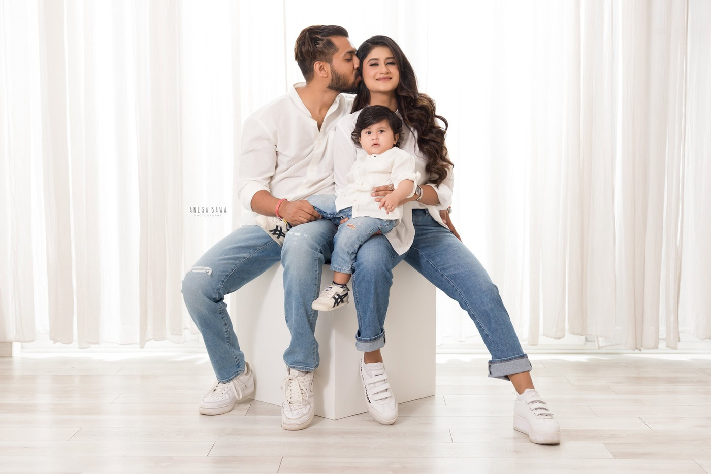 Mom, dad, and their cute son posing in a white shirt and blue denims, sitting on a white stool against a white backdrop, captured during a family photography session by Anega Bawa in Delhi, Gurgaon. Their coordinated outfits and cheerful expressions highlight the warmth of their family bond.