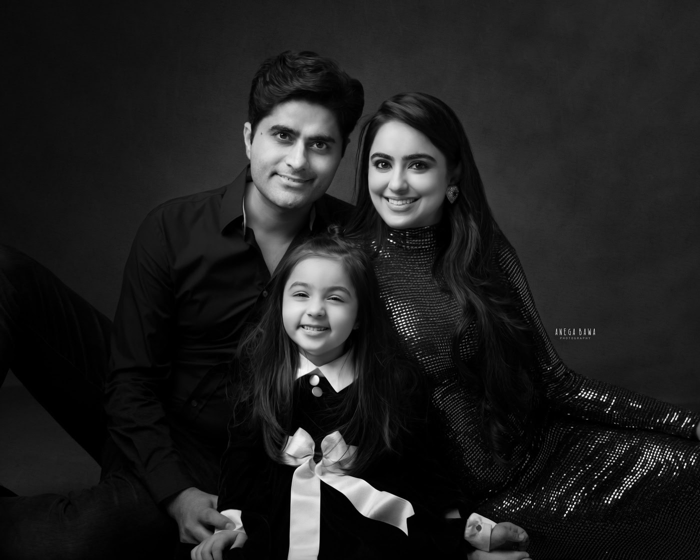 Mom, dad, and daughter all wearing black, posing cutely against a black backdrop, captured during a family photography session by Anega Bawa in Delhi, Gurgaon. Their matching outfits and joyful expressions create a striking and cohesive family moment.