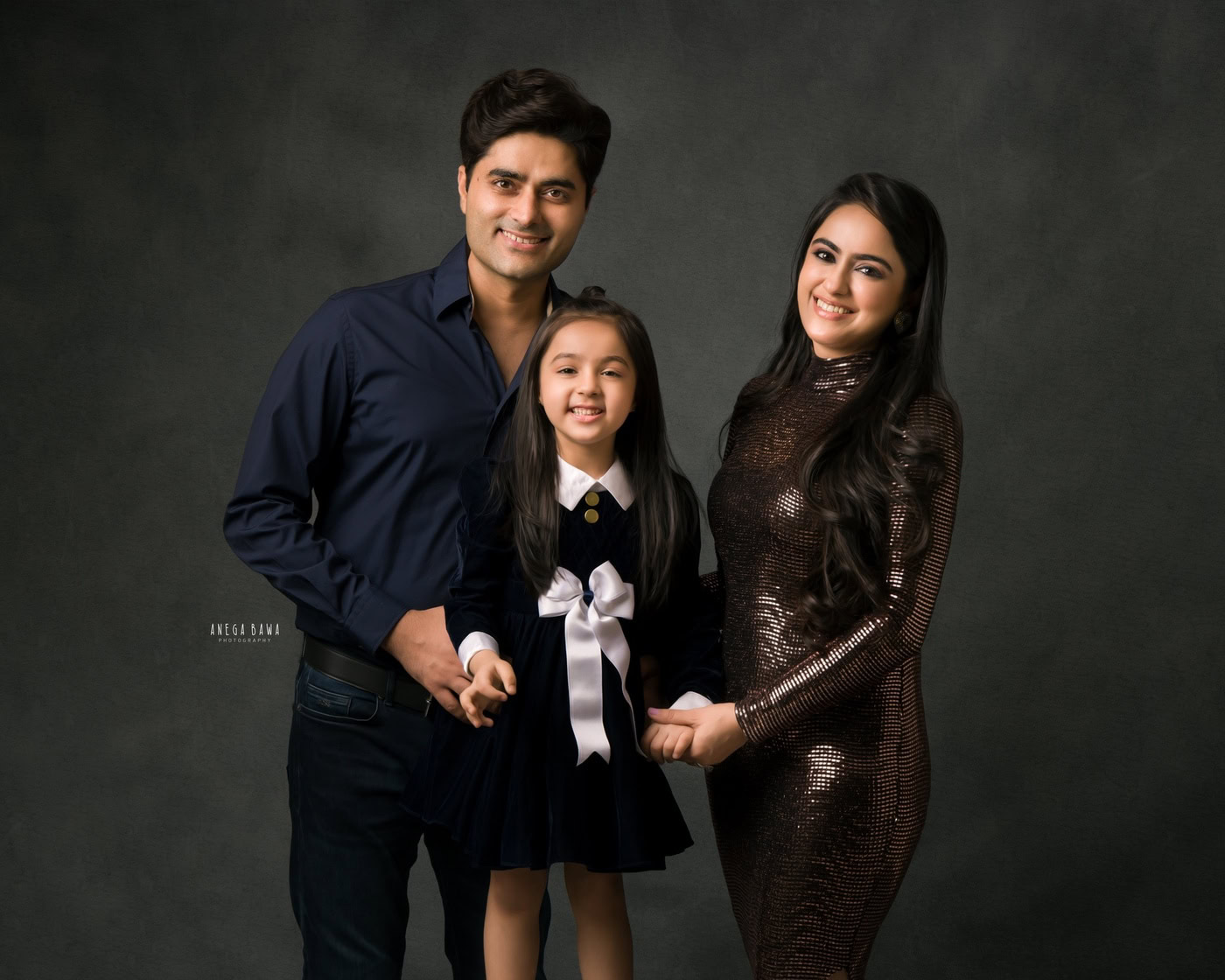Mom and dad standing, holding their daughter against a grey backdrop, captured during a family photography session by Anega Bawa in Delhi, Gurgaon. Their loving poses and smiles highlight the special bond they share as a family.