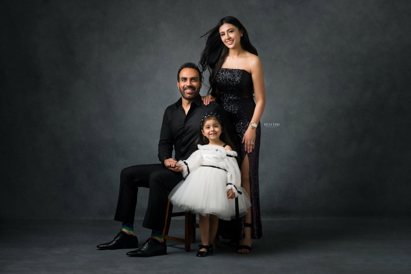 Mom, dad, and daughter all wearing white dresses, posing cutely against a grey backdrop, captured during a family photography session by Anega Bawa in Delhi, Gurgaon. Their coordinated outfits and joyful expressions create a bright and charming family moment.