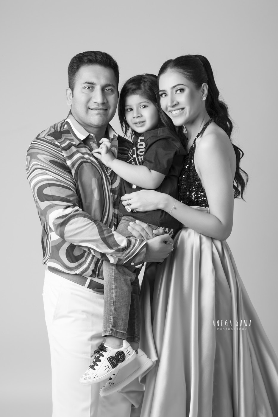 Mom and Dad holding their son in a cute pose against a black-and-white backdrop during a family photography session by Anega Bawa in Delhi, Gurgaon. The family's joy and love are beautifully captured in this timeless moment.