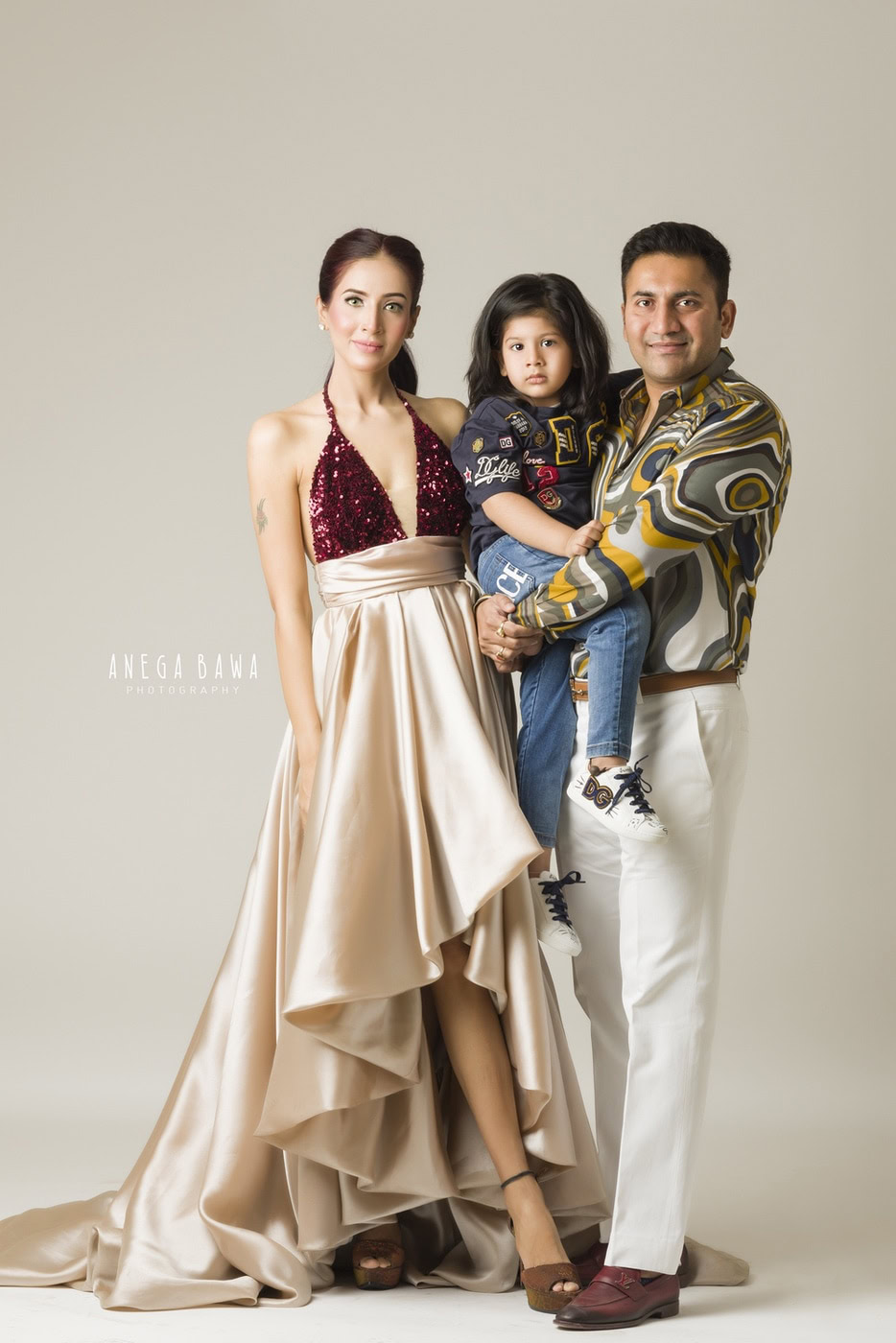 Mom and Dad holding their son in a printed shirt against a light beige backdrop during a family photography session by Anega Bawa in Delhi, Gurgaon. The warmth and affection of the family shine through in this delightful capture.