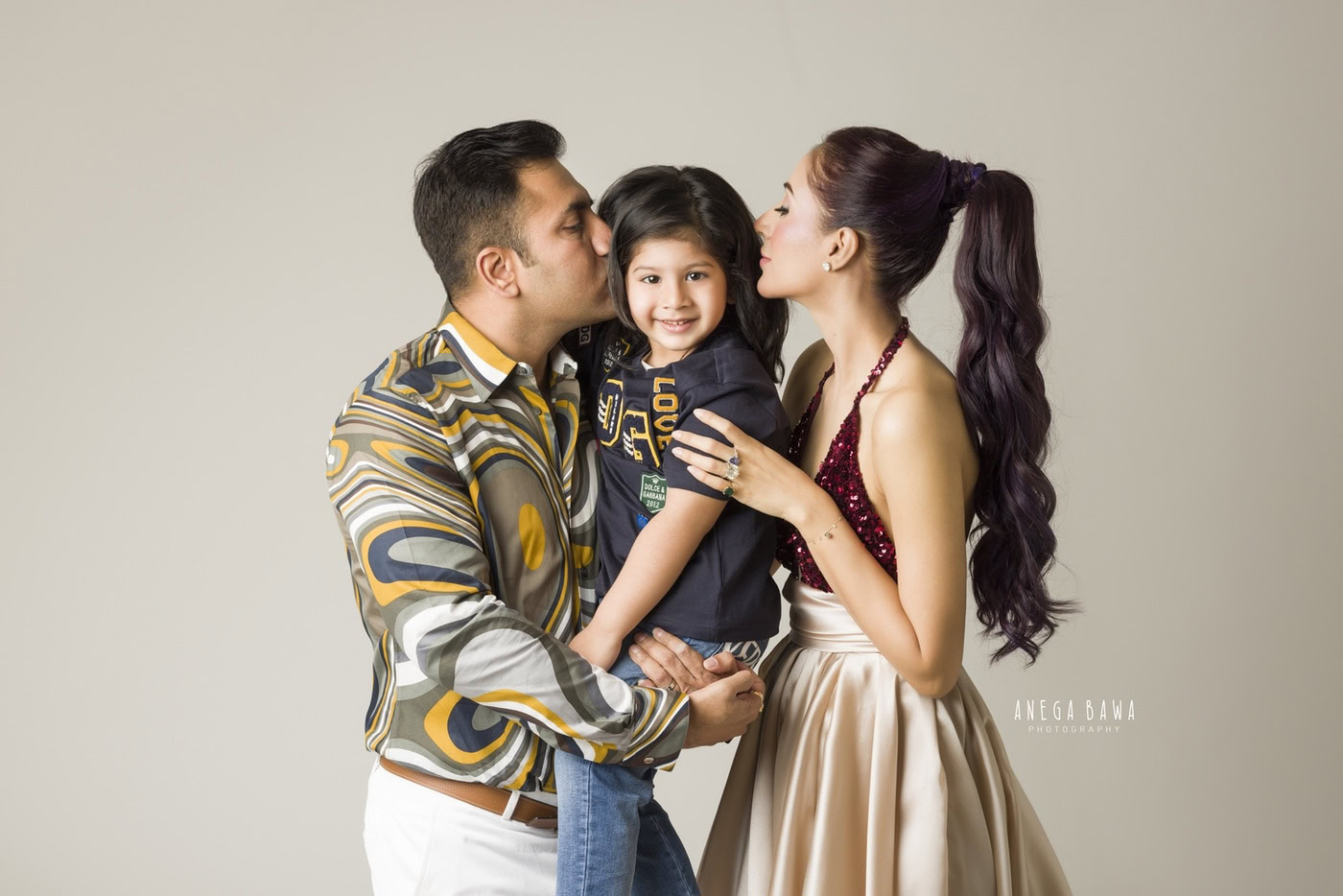 Mom and Dad kissing their son in a cute pose against a light beige backdrop during a family photography session by Anega Bawa in Delhi, Gurgaon. This heartwarming moment showcases the love and joy of family togetherness.