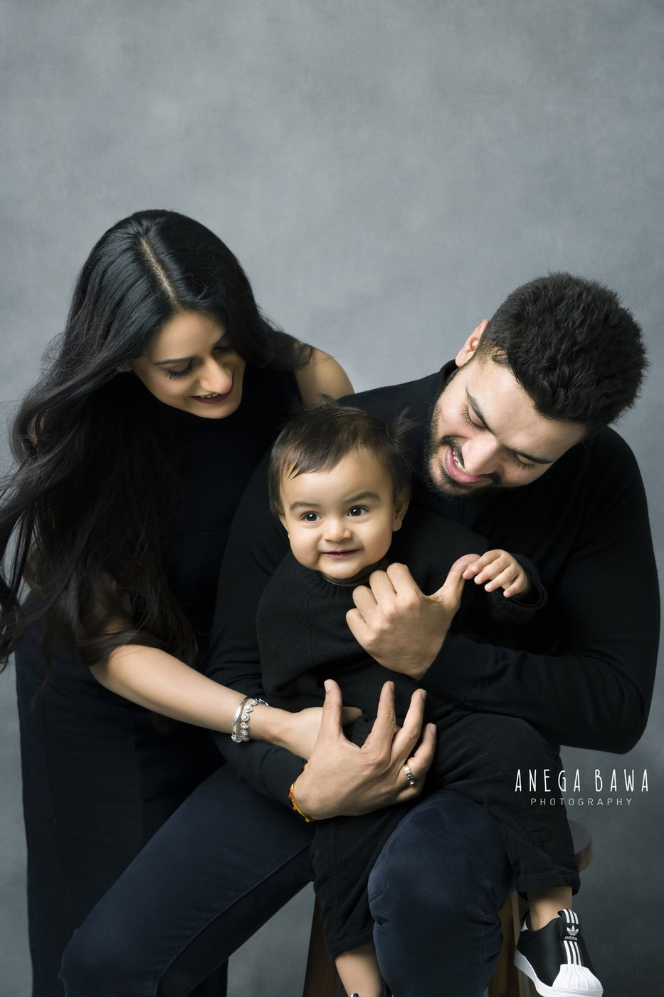 Mom, dad, and their son all wearing black, playing together in a cute smiling pose against a grey backdrop, captured during a family photography session by Anega Bawa in Delhi, Gurgaon. Their joyful interactions highlight the warmth and fun of family life.