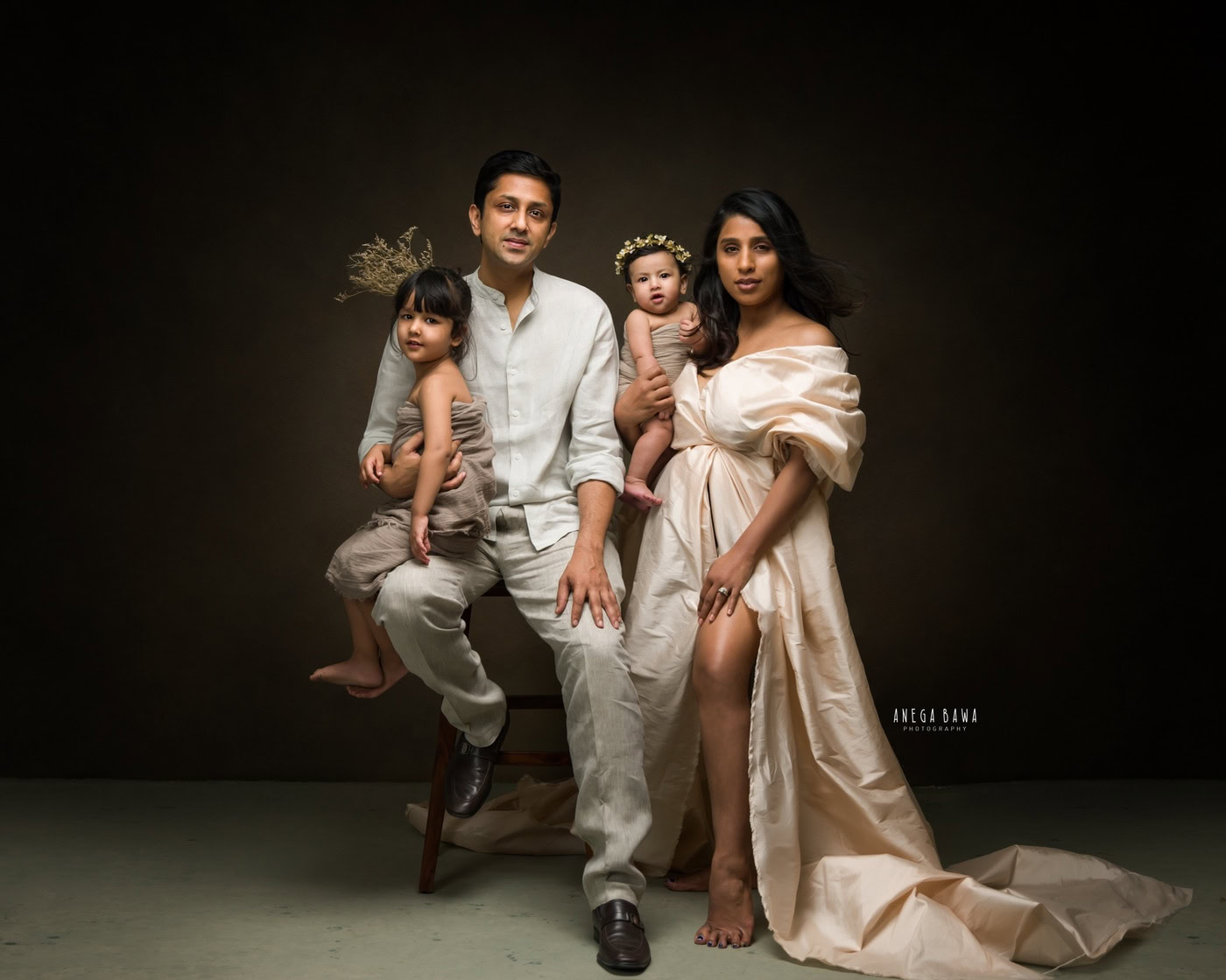 Mom and dad posing with their cute daughters, both wearing beige wraps and tiara bands against a brown backdrop, captured during a family photography session by Anega Bawa in Delhi, Gurgaon. Their coordinated outfits and joyful expressions highlight the warmth and love of their family.