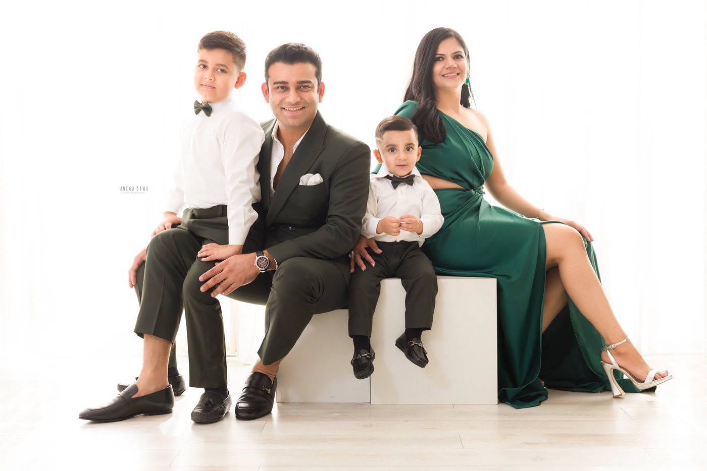 Family portrait of a stylish Indian family with two children dressed in formal attire, sitting on white cubes in a bright, modern studio setting.
