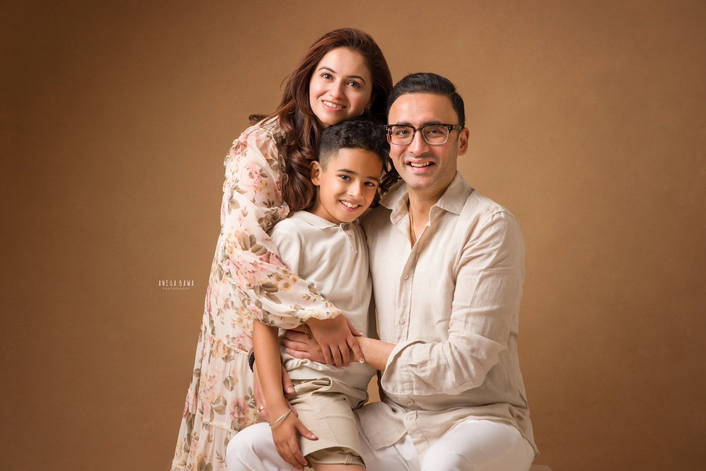 Mom, Dad, and their son posing cutely against a light brown backdrop, all smiling during a family photography session by Anega Bawa in Delhi, Gurgaon. This cheerful moment captures the essence of family joy and togetherness.