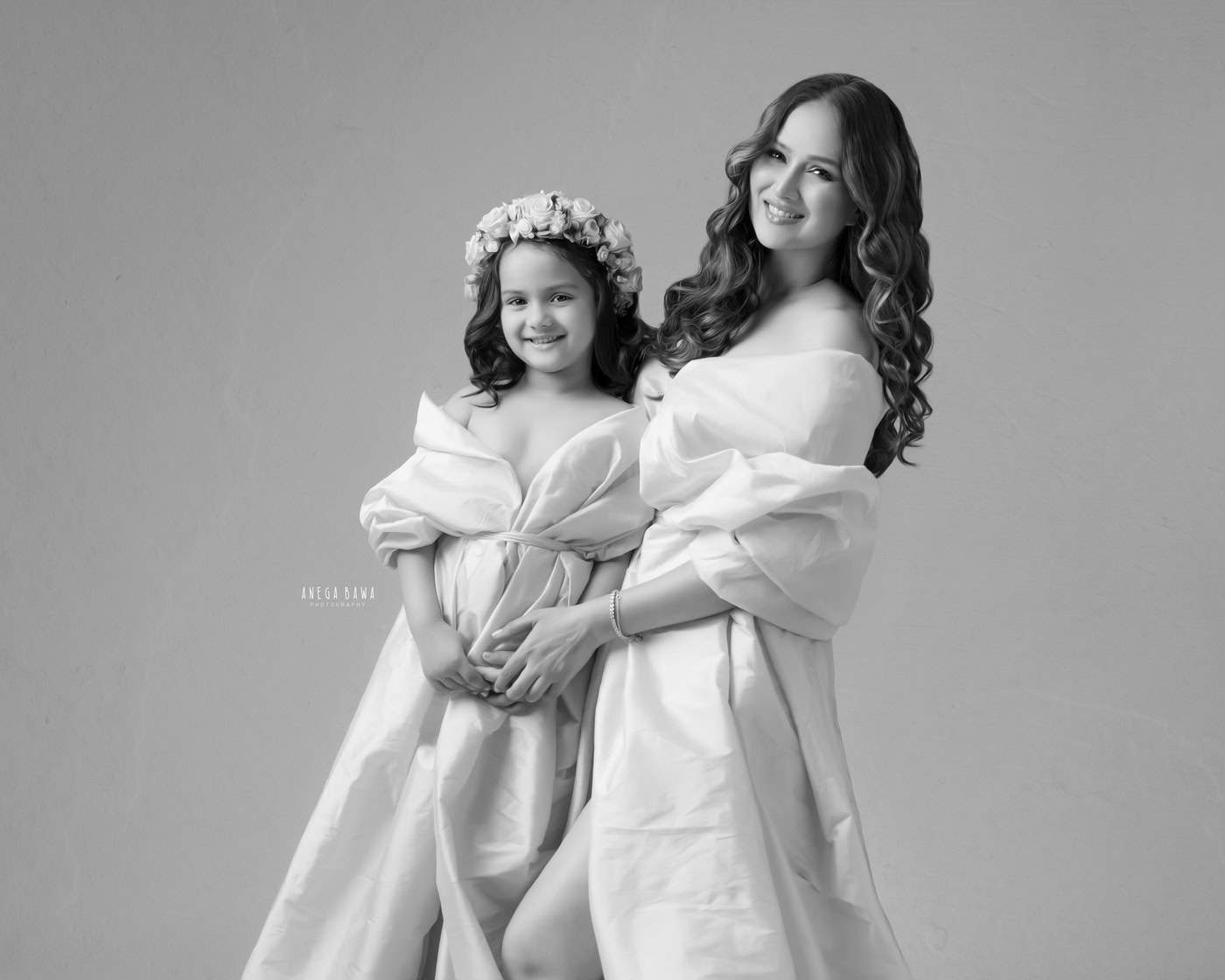 Mom and daughter posing in a white wrap dress, with the daughter wearing a tiara band, against a grey backdrop. They share a cute smiling pose, captured beautifully by Anega Bawa in Delhi, Gurgaon.