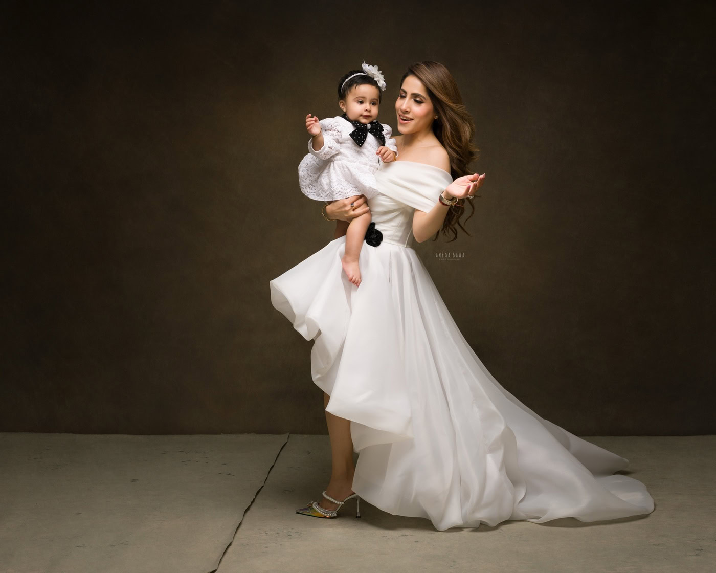 Mom and daughter posing cutely in a white dress against a brown backdrop, captured during a family photography session by Anega Bawa in Delhi, Gurgaon. Their warm smiles highlight the special bond they share.
