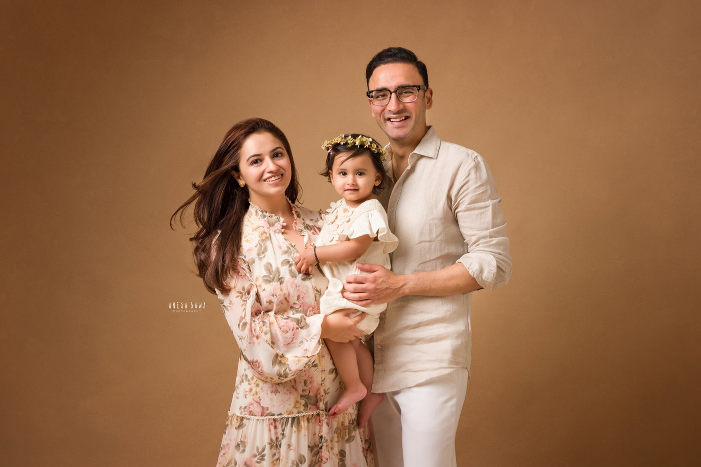 Mom holding her daughter, who is wearing a white dress and a cute tiara band, posing with Dad against a light brown backdrop. Family photography by Anega Bawa in Delhi, Gurgaon, capturing precious family moments.