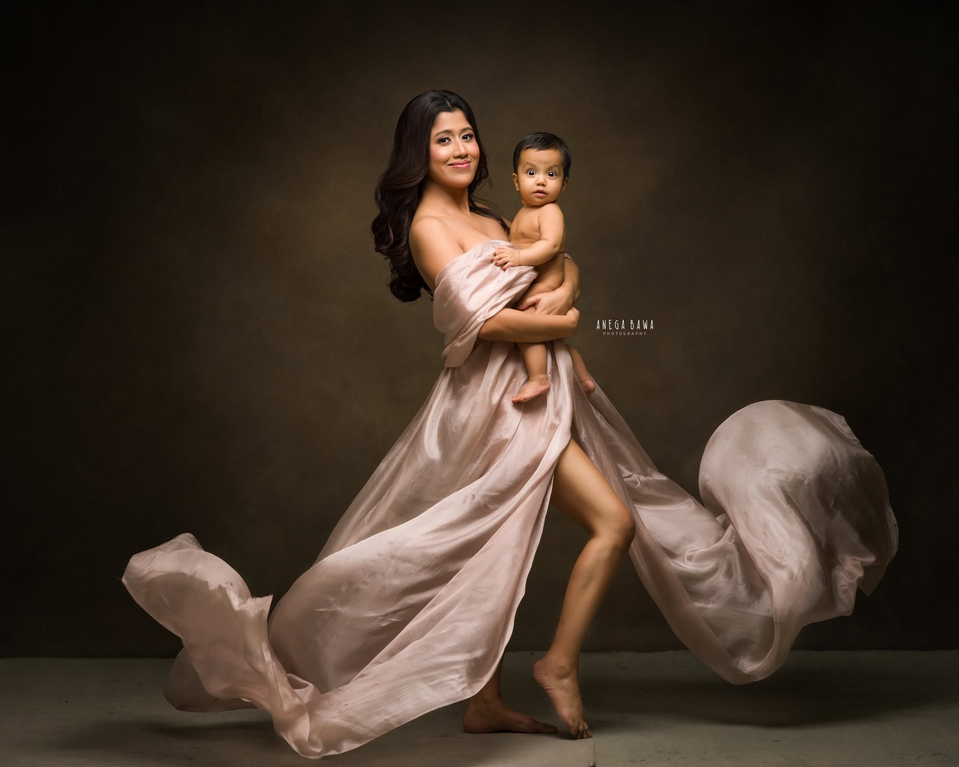 Mom in a grey wrap holding her son in a cute smiling pose against a brown backdrop, captured during a family photography session by Anega Bawa in Delhi, Gurgaon. Their joyful expressions emphasize the warmth of their bond.