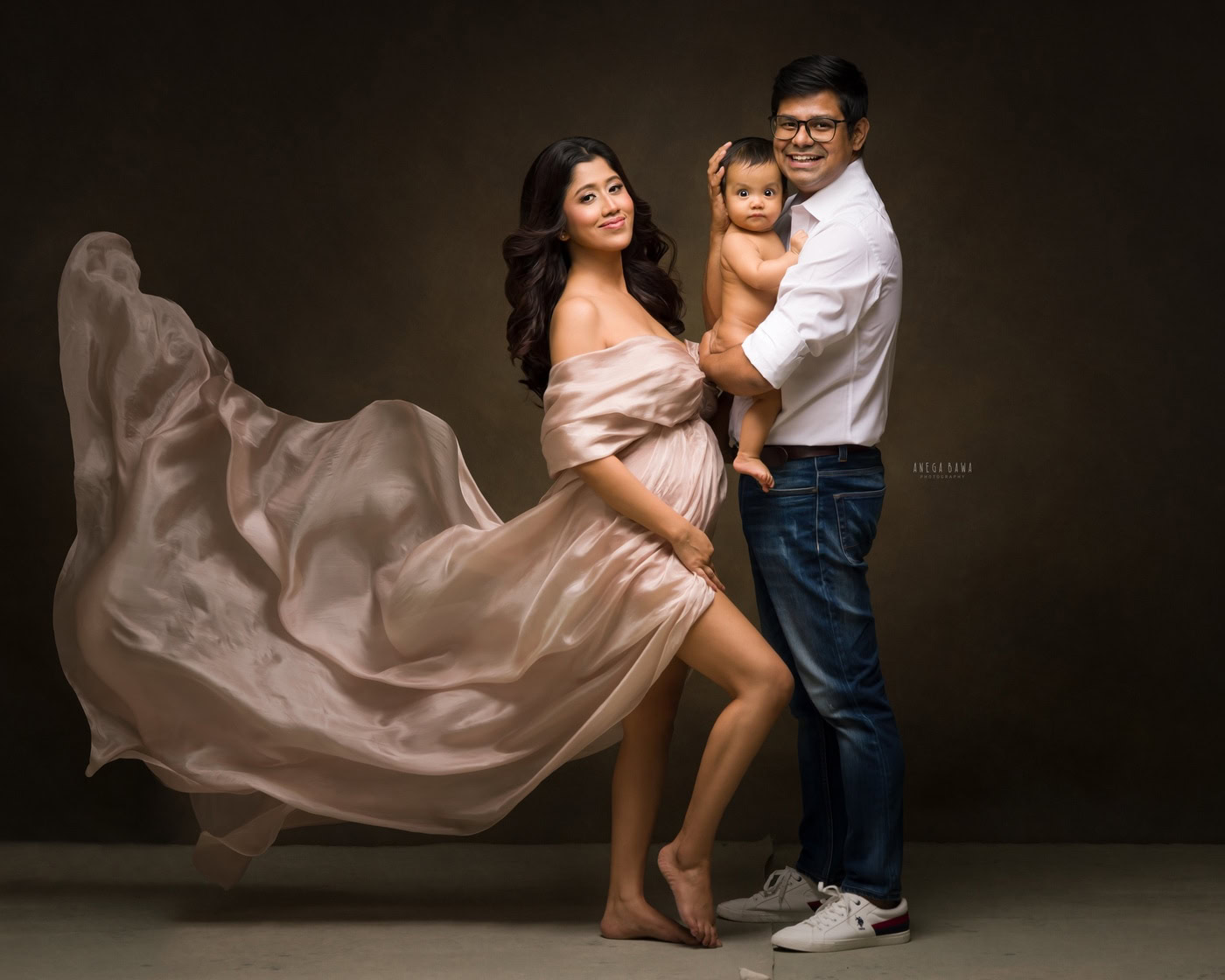 Mom posing in a beige wrap while dad holds their son in his arms against a brown backdrop in a cute smiling pose, captured during a family photography session by Anega Bawa in Delhi, Gurgaon. Their joyful expressions showcase the love and connection of their family.