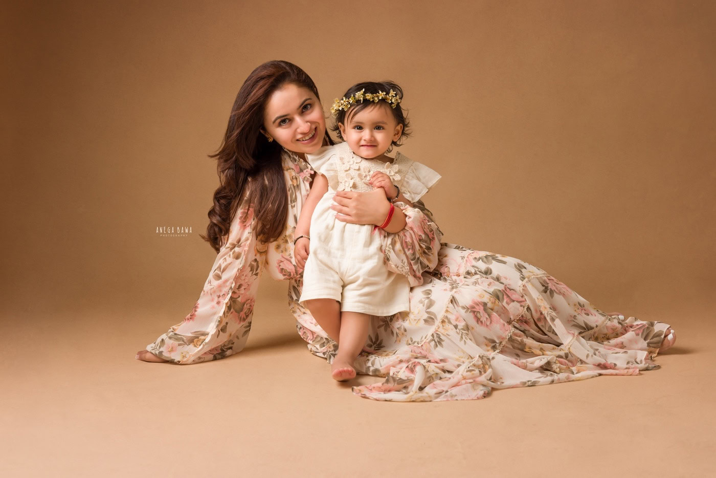 Mom posing with her cute daughter lying on the floor, both sharing a smiling pose against a light brown backdrop in family photography by Anega Bawa, Delhi, Gurgaon.