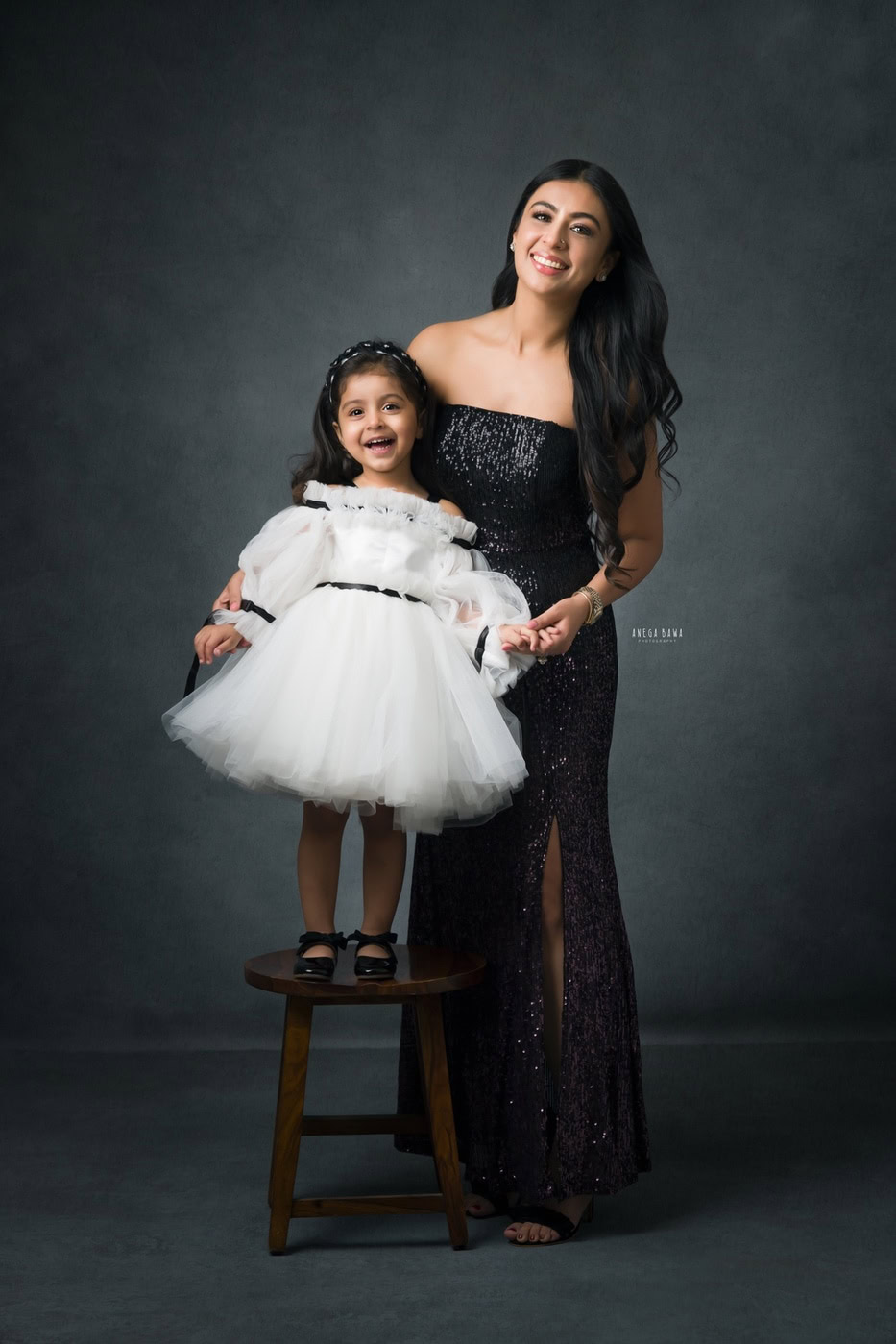 Mom posing with her cute daughter standing on a stool and wearing a white dress against a grey backdrop, captured during a family photography session by Anega Bawa in Delhi, Gurgaon. Their playful interaction highlights the joy and love of their bond.
