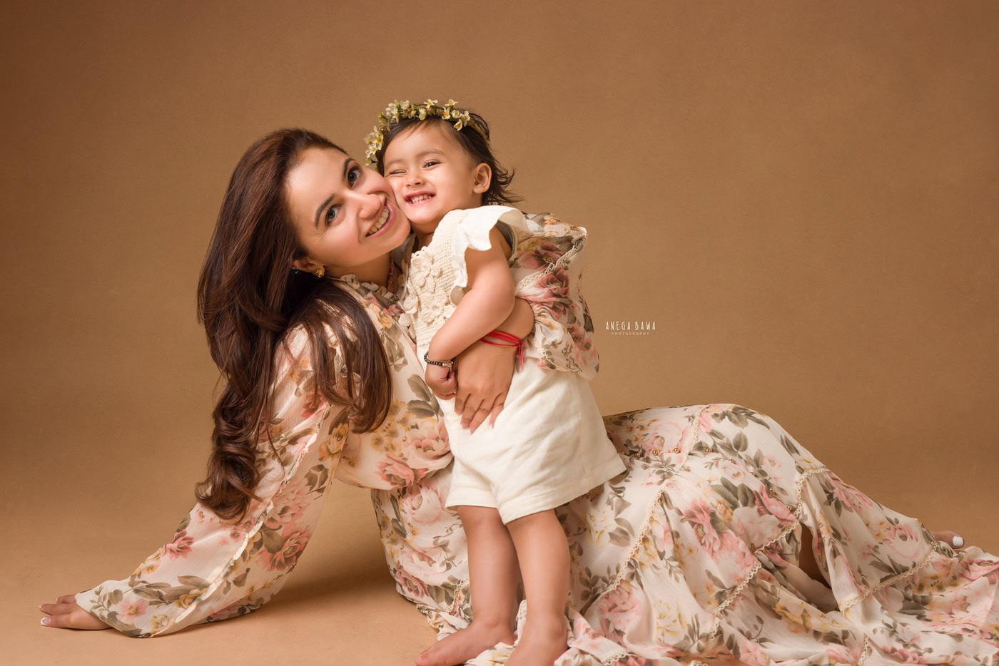 Mom posing with her cute daughter, who is wearing a white dress and a tiara band, against a light brown backdrop in family photography by Anega Bawa, Delhi, Gurgaon.
