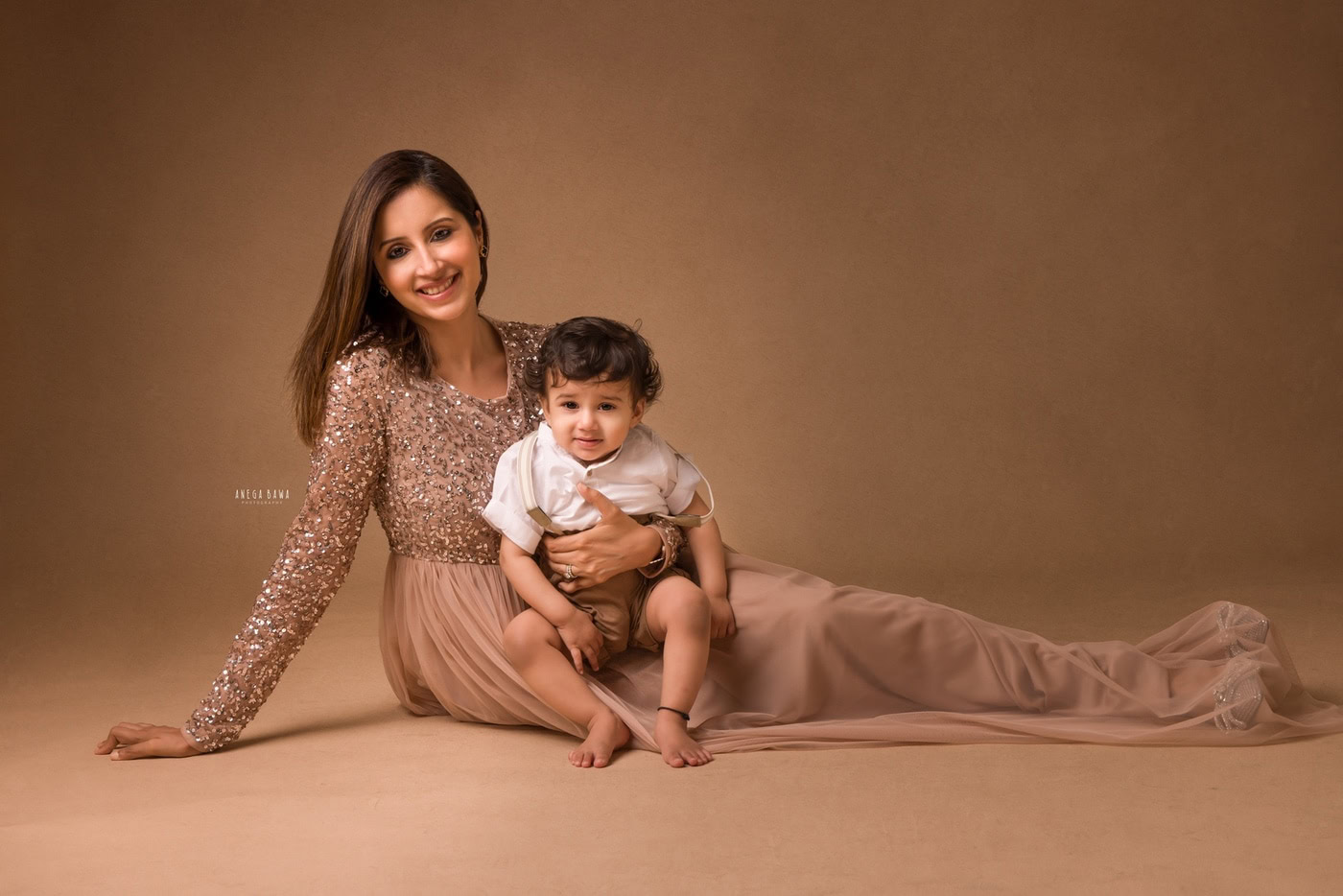 Mom posing with her son sitting on the floor against a beige backdrop, captured during a family photography session by Anega Bawa in Delhi, Gurgaon. Their warm smiles and close proximity showcase the love and connection they share.