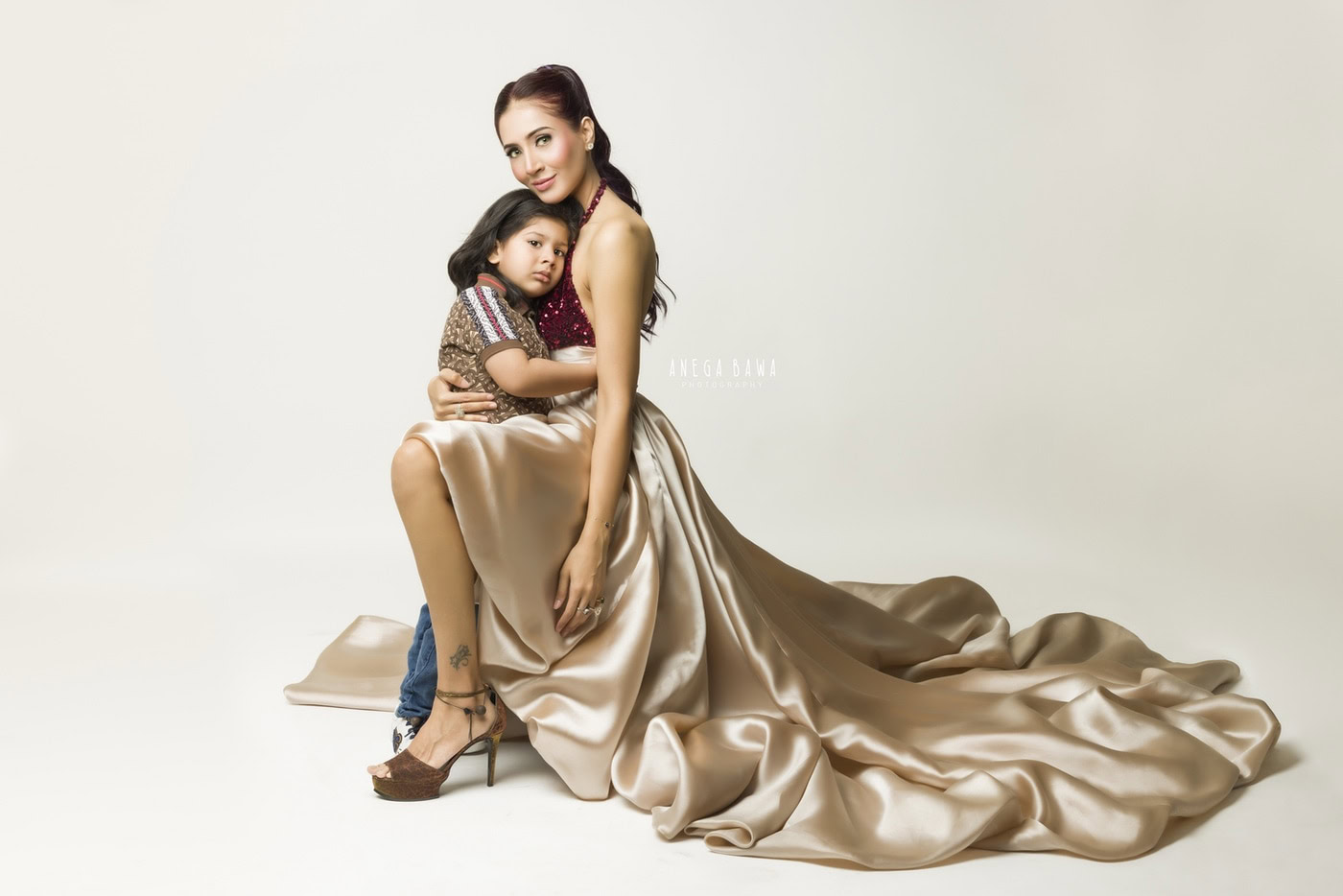 Elegant woman in a luxurious beige satin gown holding a young girl, both posing on a white studio background; professional photography for family or fashion portraits.