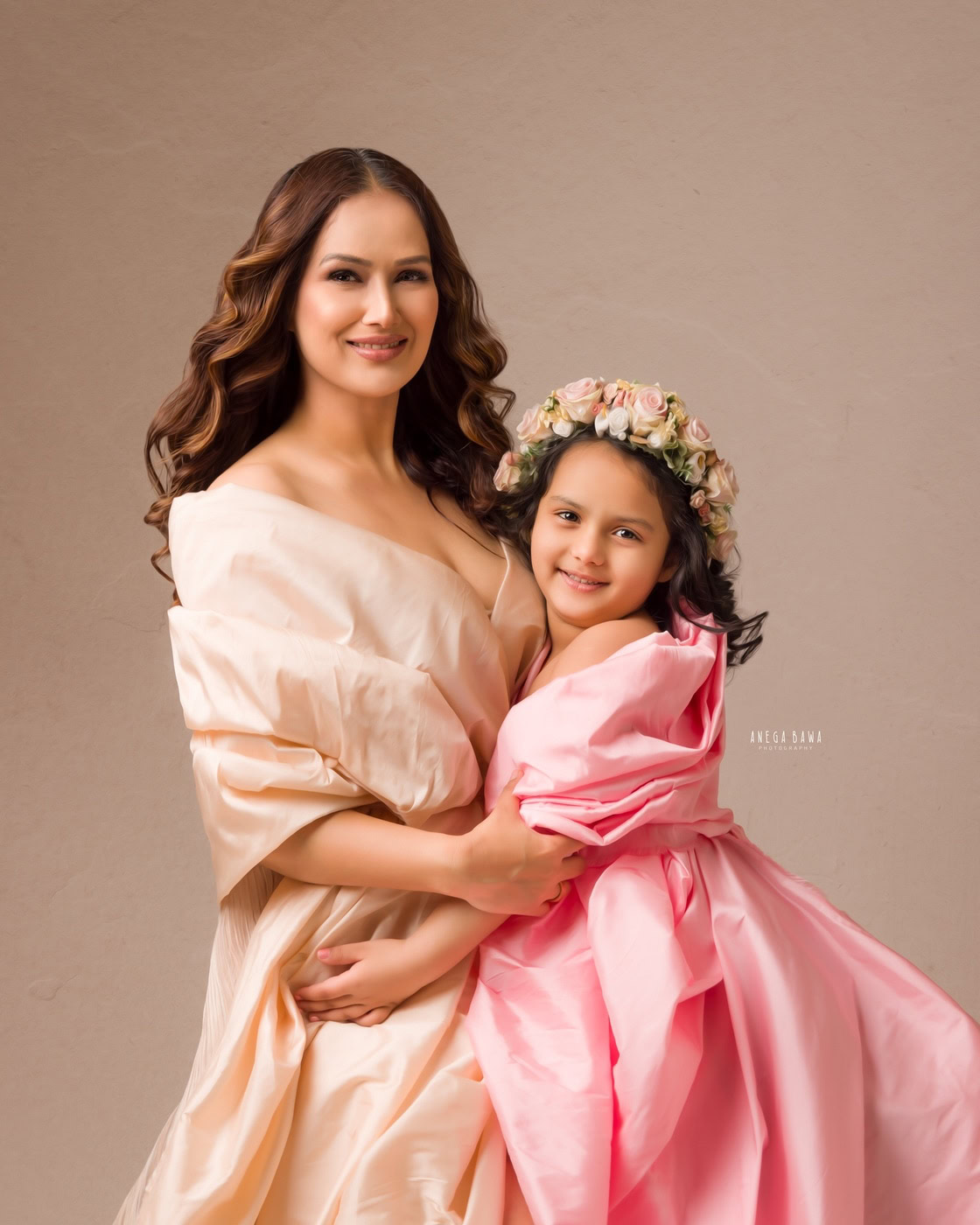 Mom in a beige wrap dress poses with her daughter, who wears a pink wrap and a cute floral tiara band, against a beige backdrop. Captured beautifully in family photography by Anega Bawa in Delhi, Gurgaon.