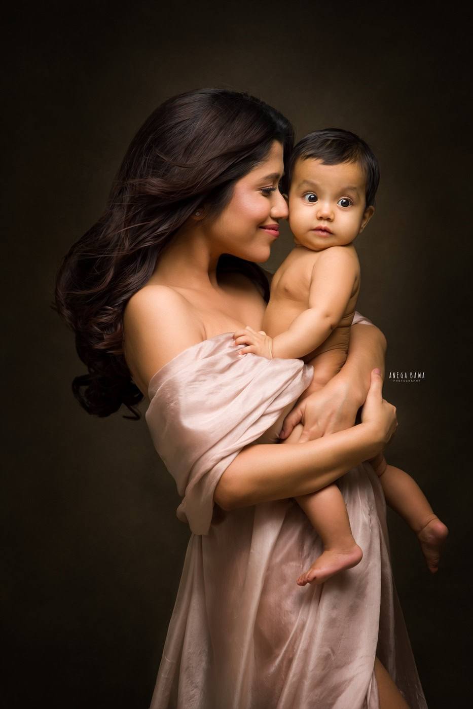 Mom wearing a grey wrap dress, cutely holding her son against a brown backdrop, captured during a family photography session by Anega Bawa in Delhi, Gurgaon. Their warm smiles and loving poses highlight the special connection they share.