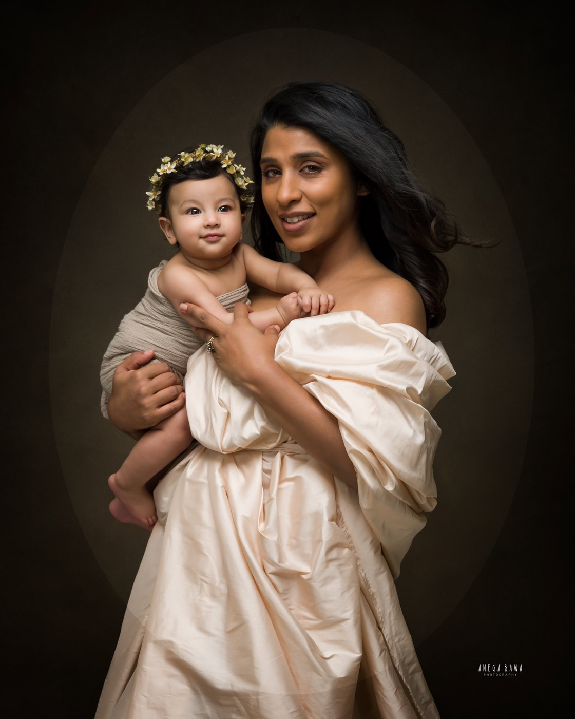 Mom wearing a white wrap dress, holding her cute daughter who is wearing a tiara band, against a brown backdrop, captured during a family photography session by Anega Bawa in Delhi, Gurgaon. Their loving poses and cheerful expressions highlight the special bond they share.
