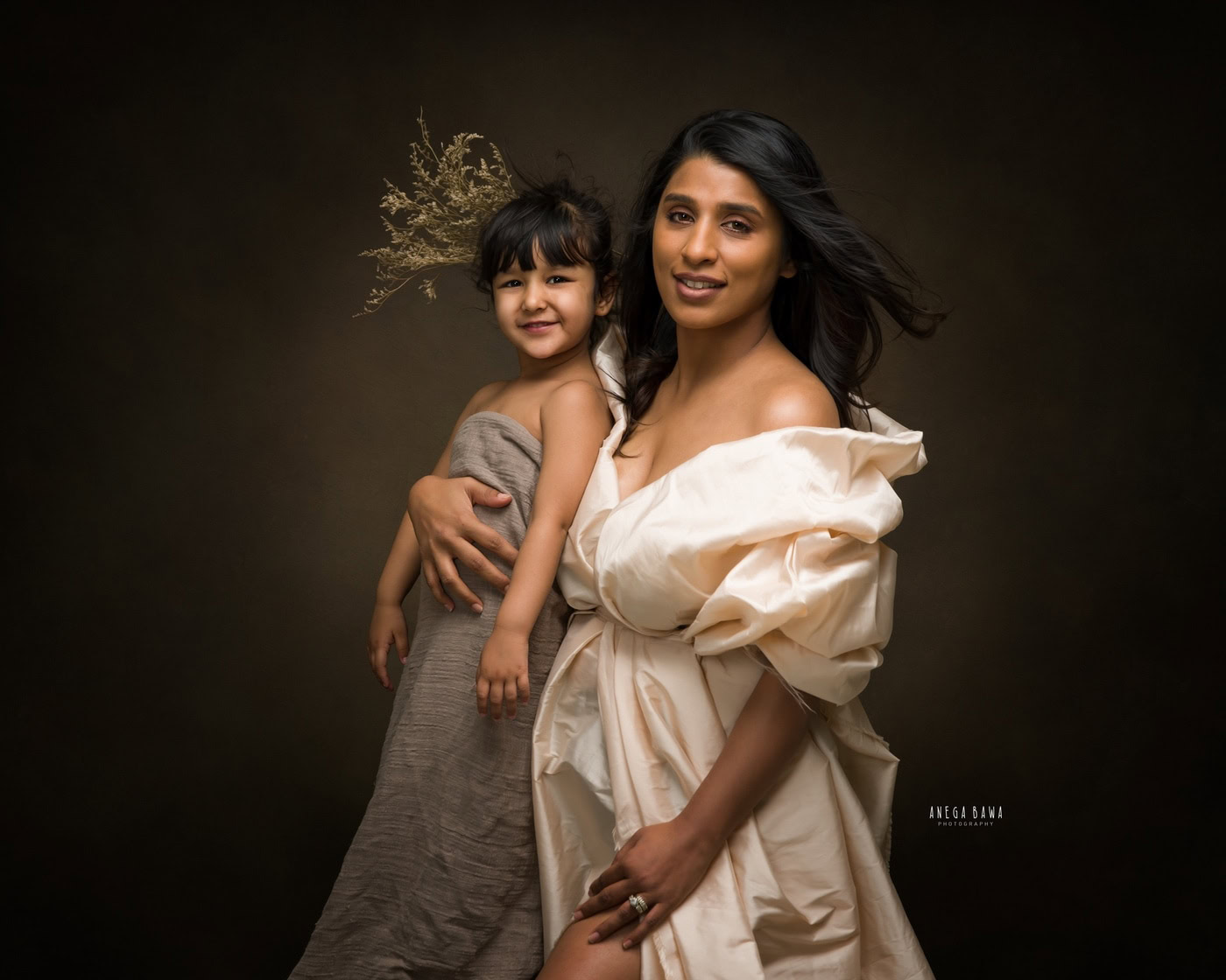 Mom wearing a white wrap dress, posing with her daughter in a grey wrap and a beautiful hair accessory against a brown backdrop, captured during a family photography session by Anega Bawa in Delhi, Gurgaon. Their lovely outfits and joyful expressions highlight the bond they share.