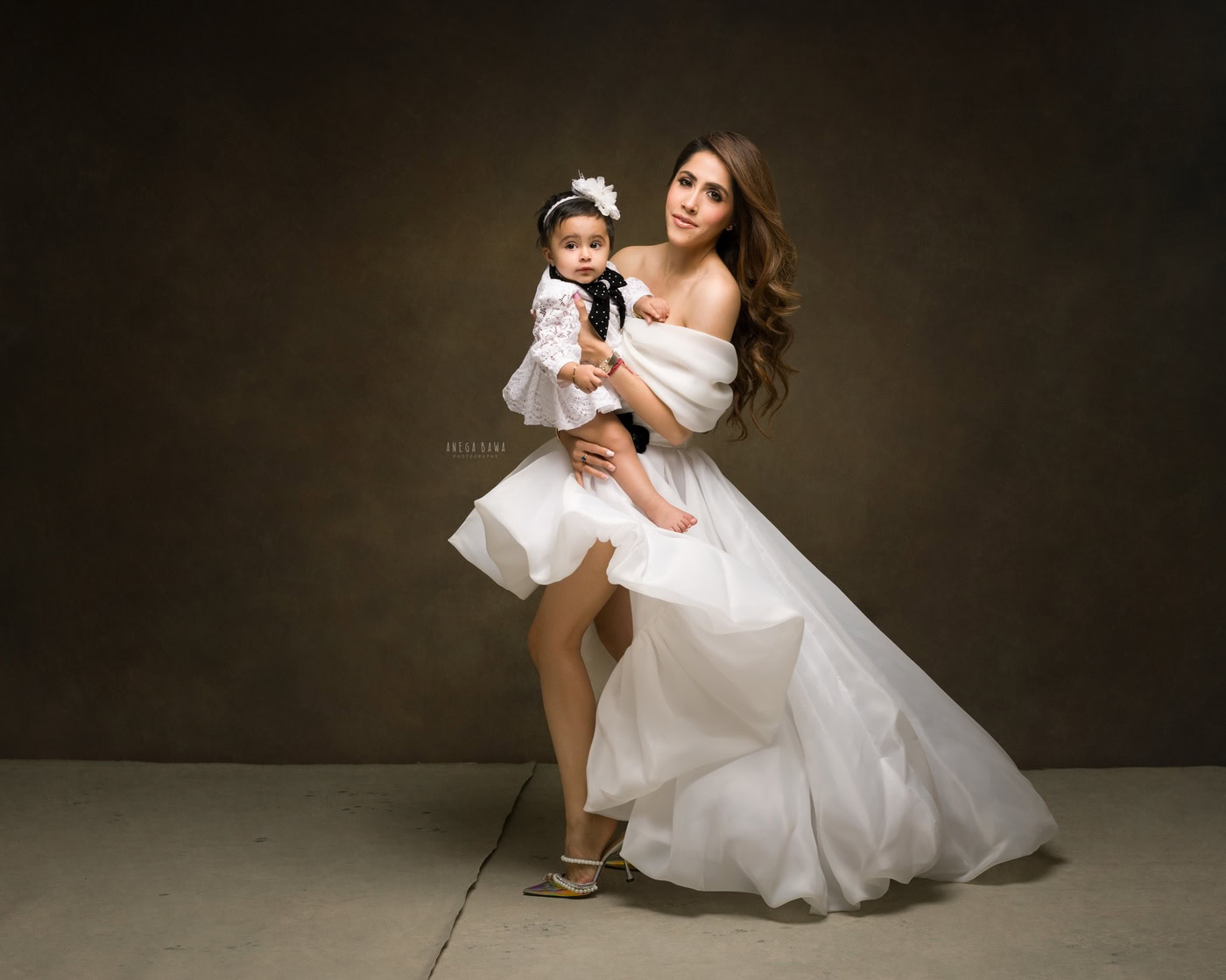Elegant woman in a white gown holding a baby girl in a lace dress with a bow headband, studio portrait, high fashion photography.