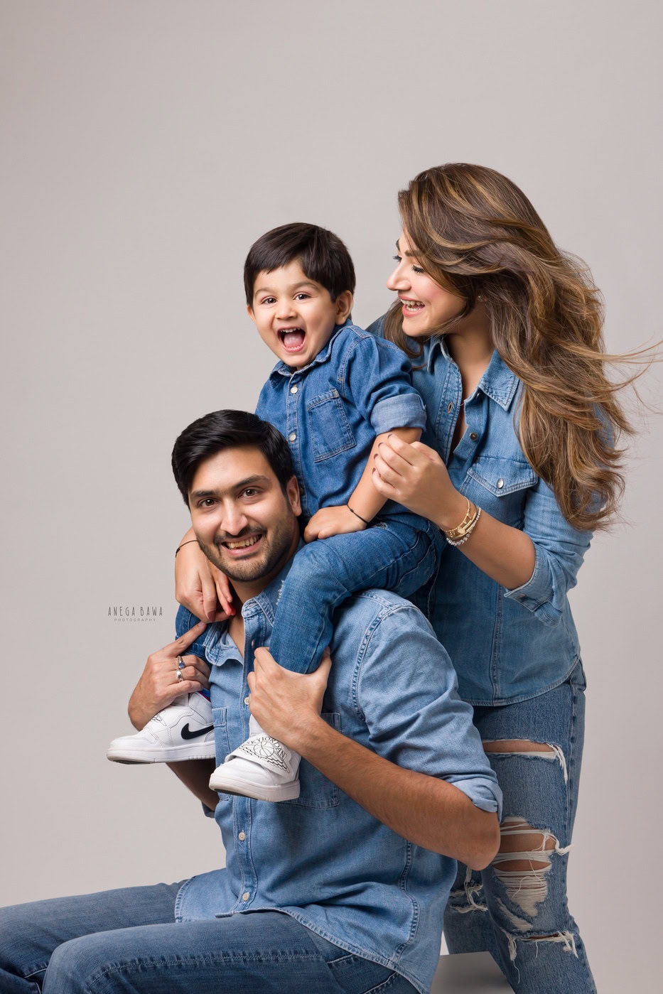 Son sitting on dad's shoulders, with mom posing alongside them, all wearing denim shirts in a cute smiling pose, captured during a family photography session by Anega Bawa in Delhi, Gurgaon. Their joyful expressions highlight the fun and love of their family connection.