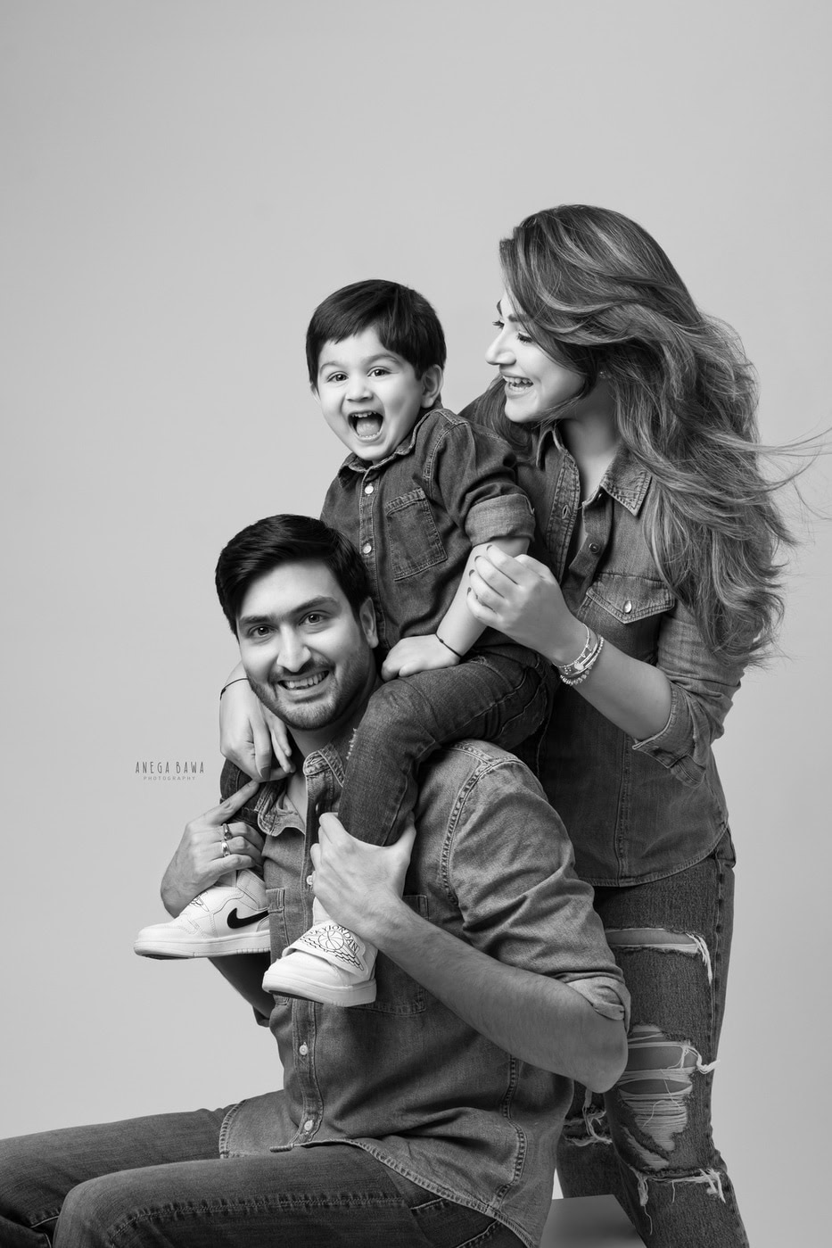 Son sitting on dad's shoulders, with mom posing in a cute smiling pose against a grey backdrop in black-and-white, captured during a family photography session by Anega Bawa in Delhi, Gurgaon. Their joyful expressions beautifully capture the warmth and love of their family.