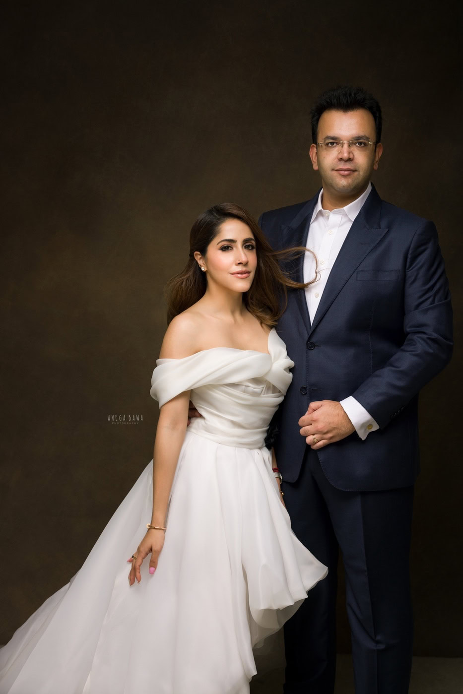 Wife wearing a white dress, posing with her husband in a cute pose against a brown backdrop, captured during a family photography session by Anega Bawa in Delhi, Gurgaon. Their cheerful expressions and coordinated outfits showcase the love and joy in their relationship.