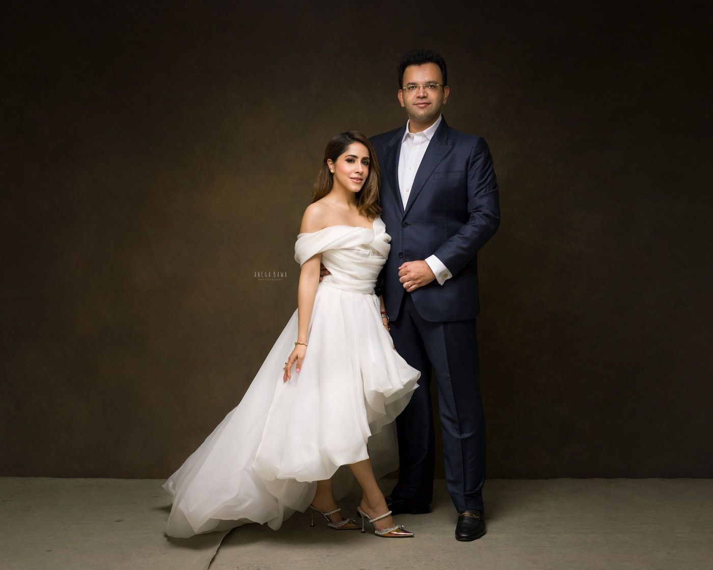 Wife wearing a white gown, posing with her husband against a brown backdrop, captured during a family photography session by Anega Bawa in Delhi, Gurgaon. Their elegant outfits and warm smiles beautifully highlight the affection in their relationship.