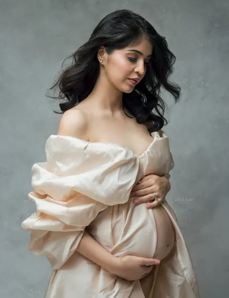 Expecting mother in an off-shoulder champagne satin robe with large puffed sleeves, looking down, hands under her bare bump, soft grey