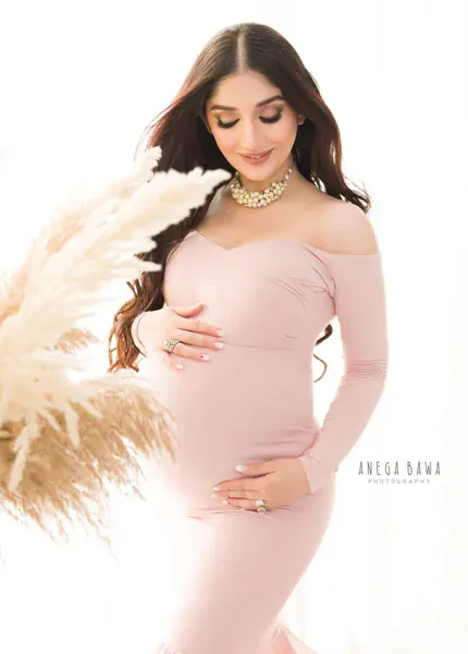 Smiling expecting mother in a blush pink off-shoulder long-sleeve fitted gown with a gold necklace, holding pampas grass in the foreground