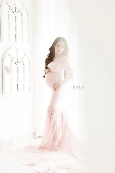 Expecting mother in a pale blush off-shoulder long-sleeve mermaid gown with a long train, hand on her bump, white wall with arched windows