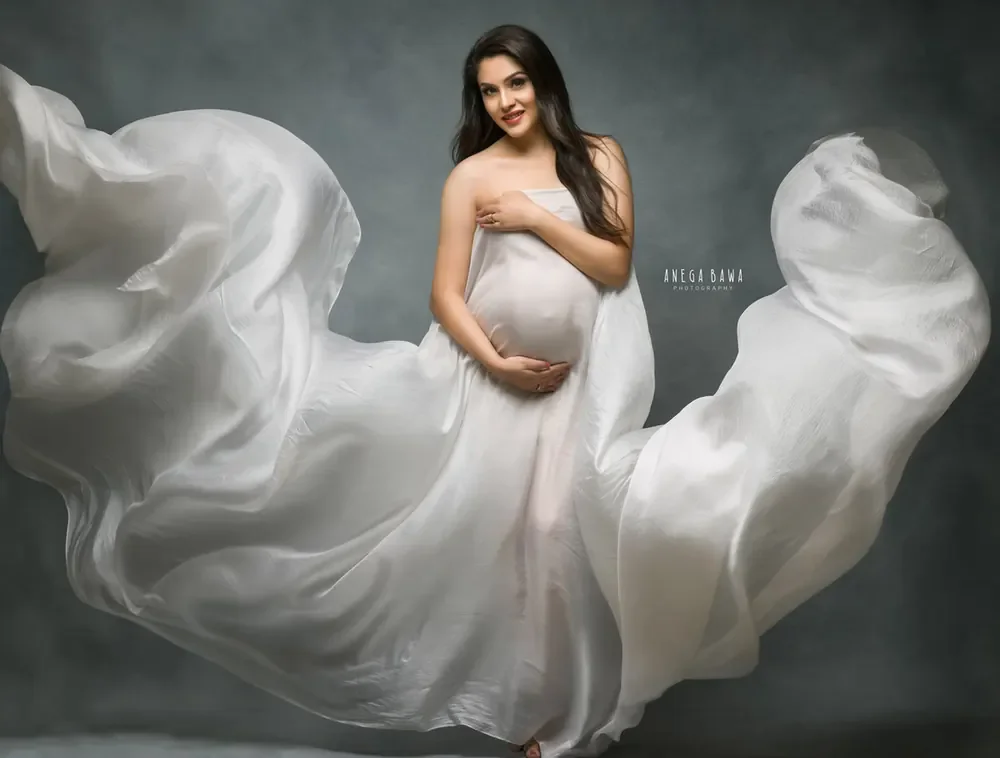 Expecting mother in a sheer white satin drape with huge billowing fabric spread around her, hands on her bump, grey-green backdrop.