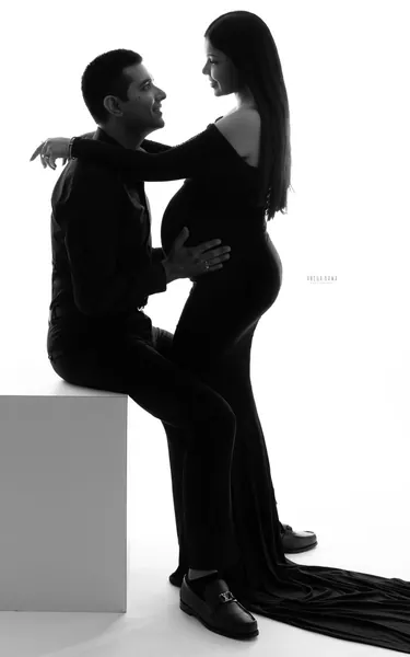 High-key black silhouette of a couple, husband seated on a white block holding his wife by the waist and bump as she stands in a fitted