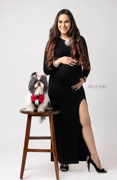 Smiling expecting mother in a black long-sleeve fitted gown with a high slit and peep-toe heels, a small shih tzu in a red bow tie on a