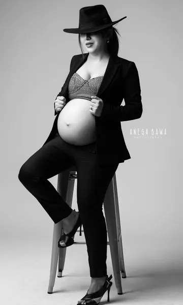 Black and white styled portrait of expecting mother seated on a stool in a black blazer, patterned bralette, trousers, heels and a