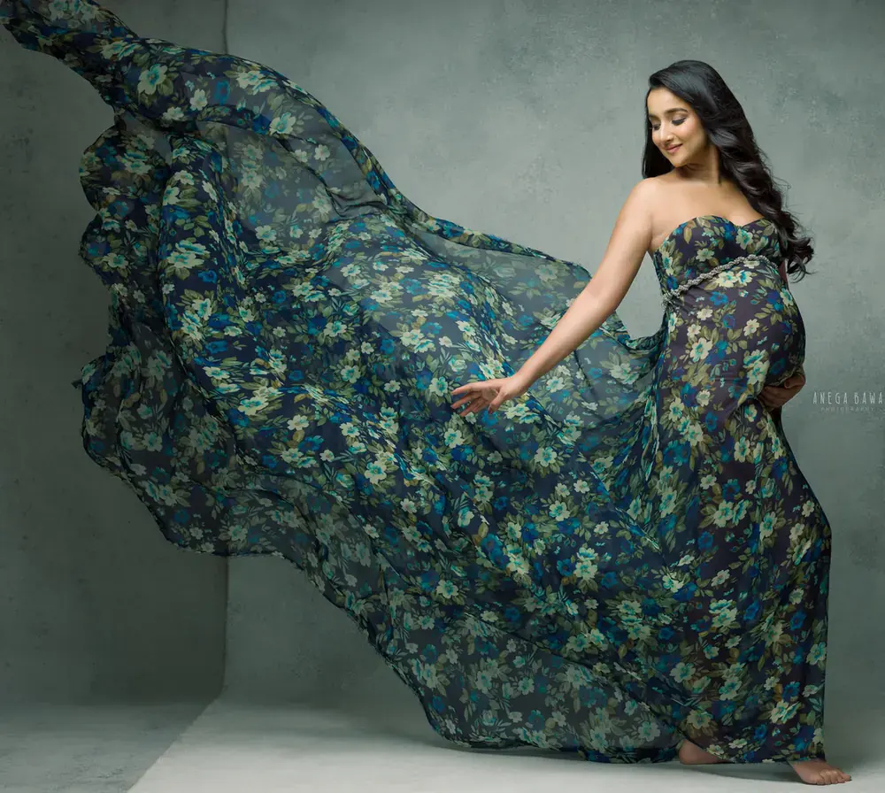 Expecting mother in a strapless blue and green floral gown with a long sheer train blowing out to one side, barefoot, hand reaching toward