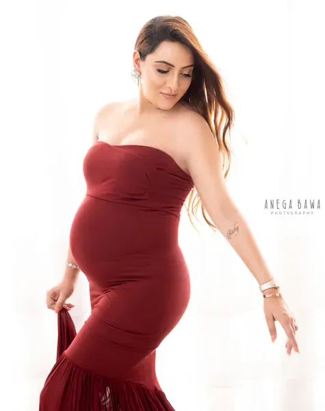 Expecting mother in a maroon strapless fitted mermaid gown looking down with one hand lifting the skirt, hair blowing, bright white