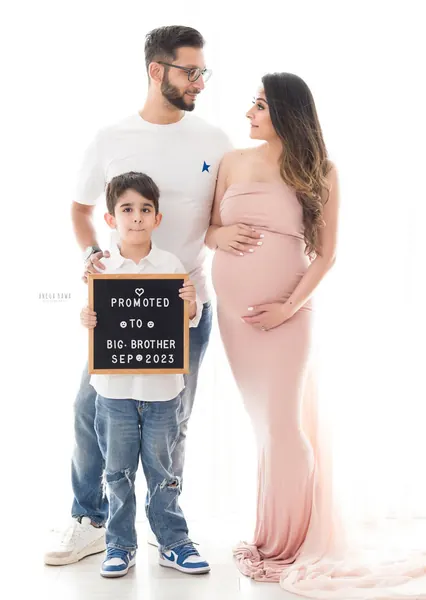 Family of three: husband in a white tee and jeans, wife in a blush pink strapless fitted gown, and their young son holding a letterboard