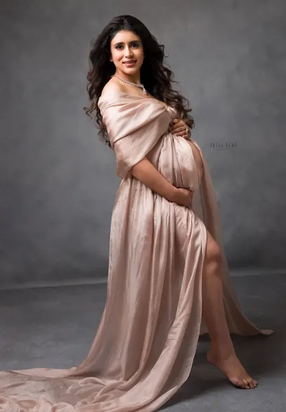 Expecting mother seated in a blush satin gown with a thigh slit and a pearl necklace, barefoot with one knee raised, hands cradling her
