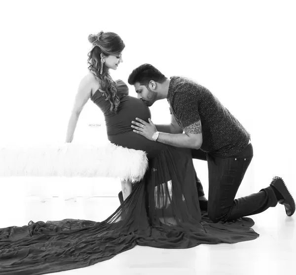High-key black and white portrait of husband kneeling to kiss the bump of his wife, who sits on a white fur bench in a strapless gown with