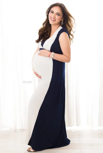 Smiling expecting mother in a colour-block navy and white sleeveless long gown, hands on her bump, bright white curtain studio.