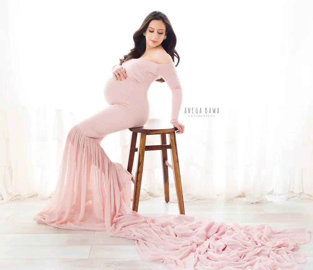 Expecting mother seated on a wooden stool in a blush pink off-shoulder long-sleeve mermaid gown with a long sheer train, hand on her bump