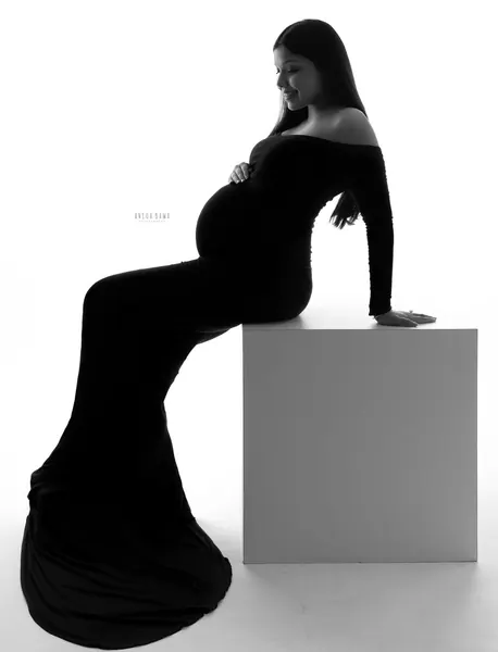 Black and white high-key silhouette of expecting mother seated on a white block in a black fitted off-shoulder long-sleeve gown with a long