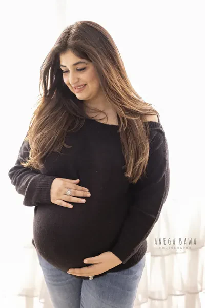 Smiling expecting mother in a dark brown knit off-shoulder sweater and jeans, long hair, looking down and cradling her bump, bright white